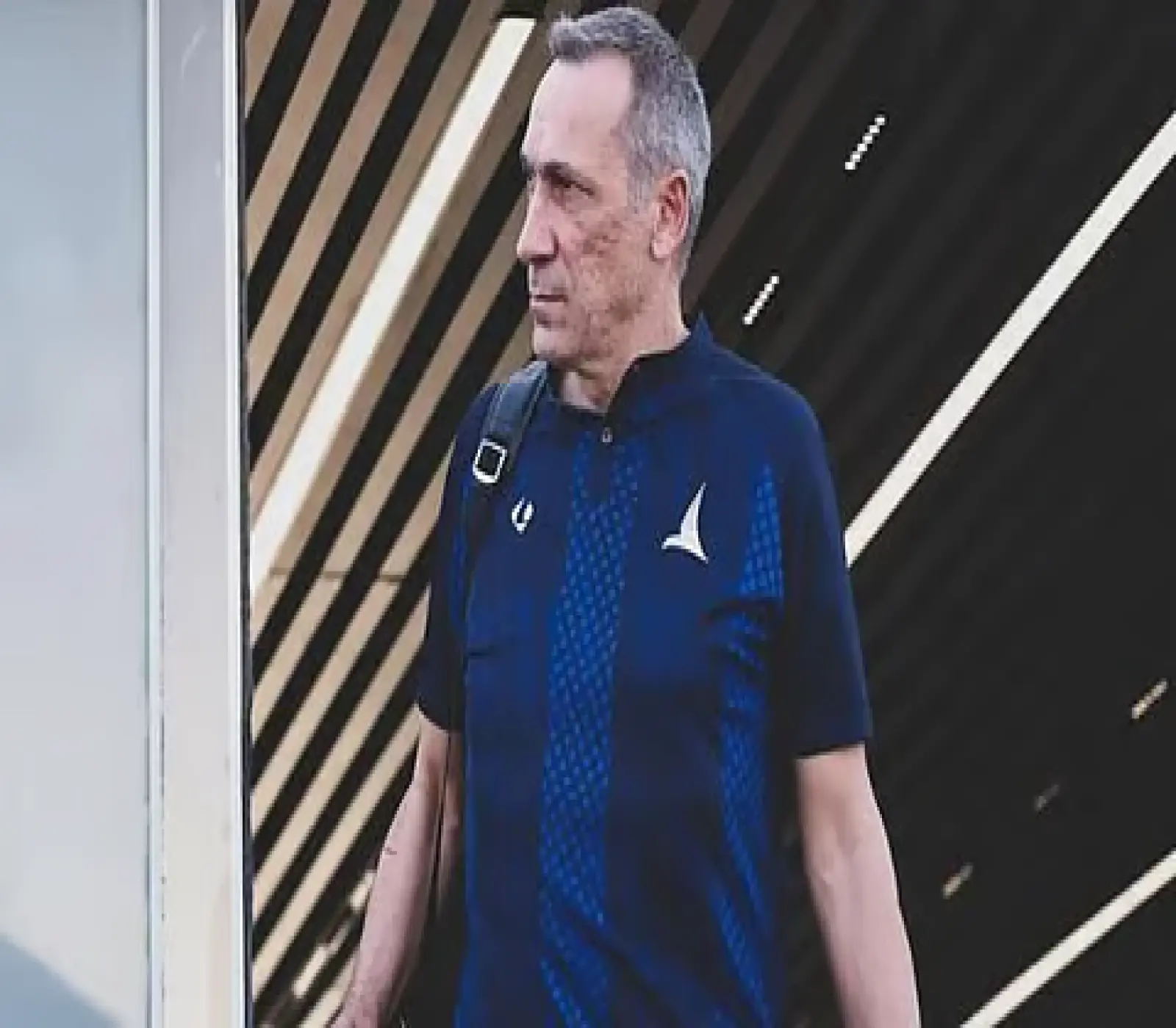 رجل يرتدي قميصاً رياضياً أزرق ويحمل حقيبة على كتفه، يسير في ممر ذو خلفية مخططة.