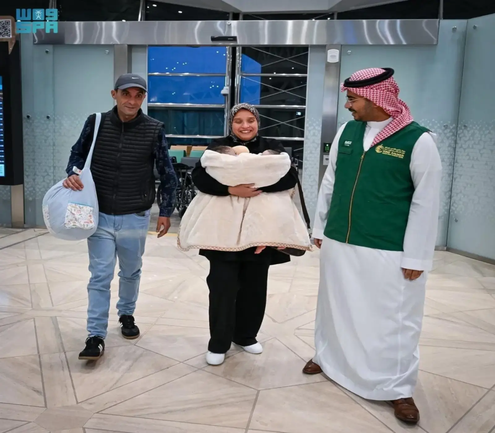 امرأة تحمل طفلاً ملفوفاً ببطانية بيضاء يرافقها رجلان داخل صالة مطار بانتظار إتمام إجراءات السفر.