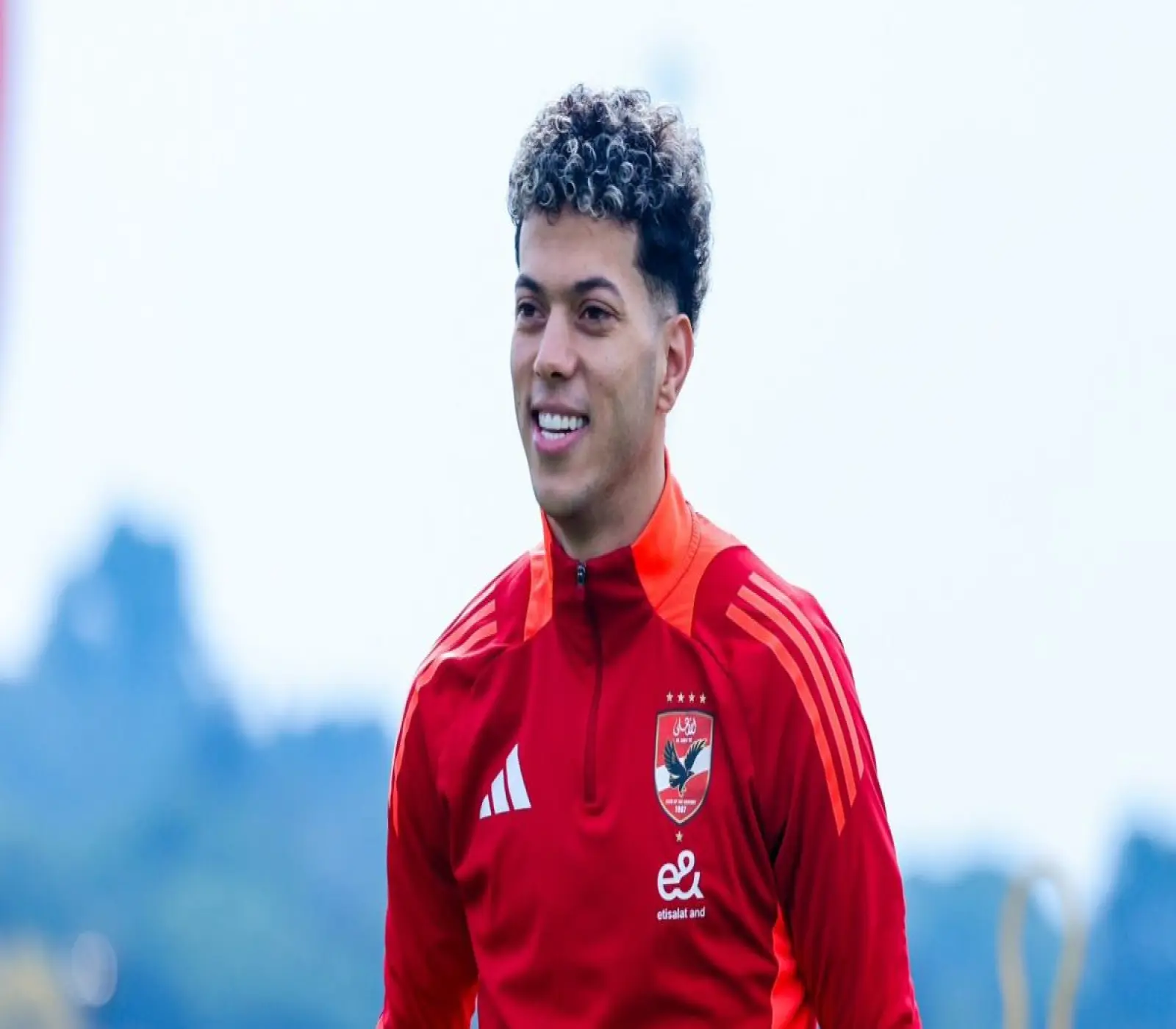 لاعب كرة القدم إمام عاشور يبتسم مرتدياً قميص التدريب الأحمر الخاص بالنادي الأهلي المصري في ملعب مفتوح.